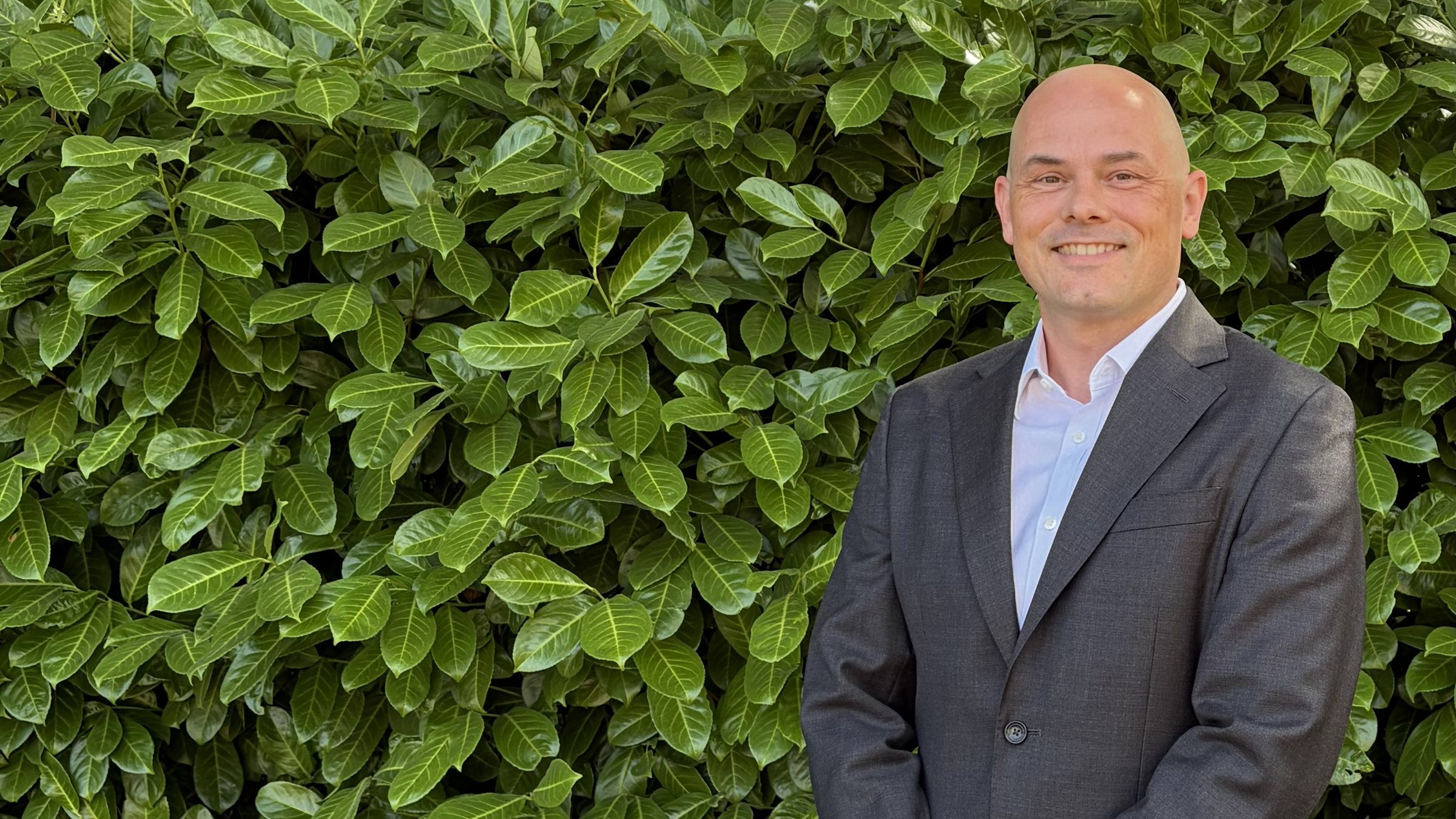 Mark Apsey in a grey suit and open-collar shirt, standing to the right of frame in front of a laurel hedge